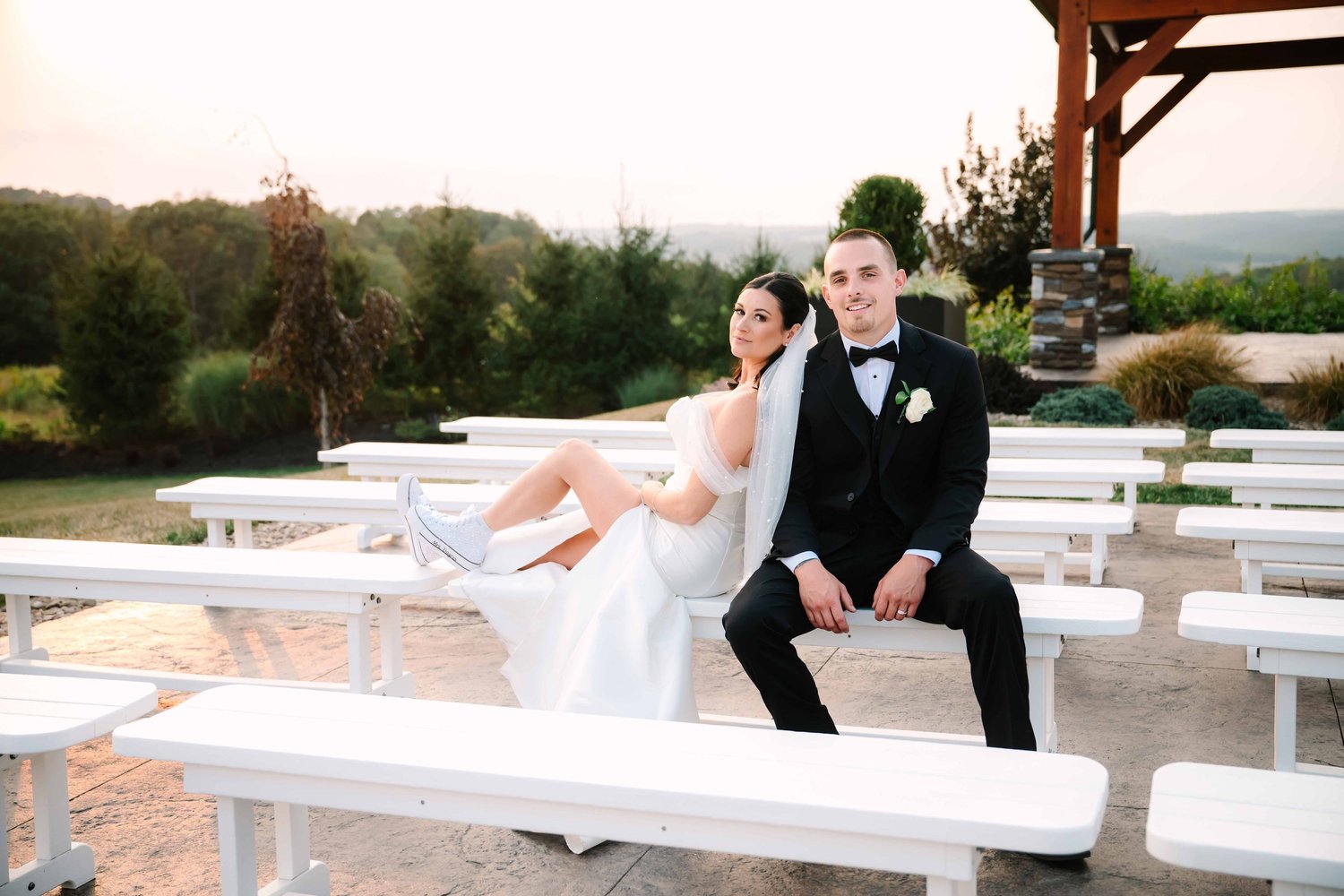 A bride in a white gown and sneakers reclines across a white bench beside her groom in a black tuxedo, on an outdoor ceremony patio at sunset